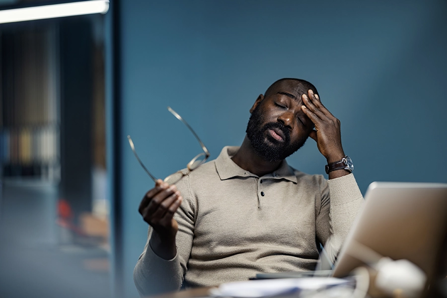 Man at his desk holding his glasses and pressing a hand to his forehead with his eyes closed.
