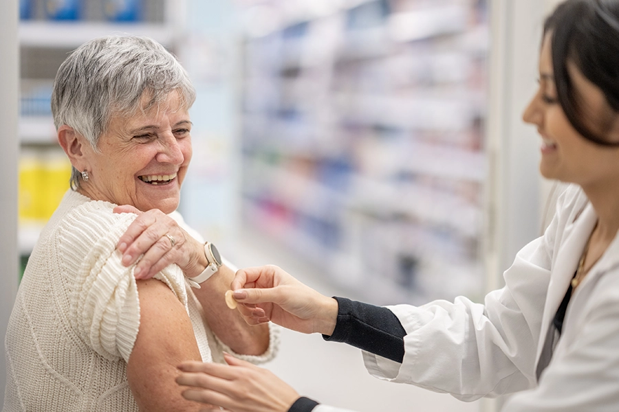 Smiling older woman in a cream sweater with her sleeve pushed up as a pharmacist in a white coat places a bandage on her arm.